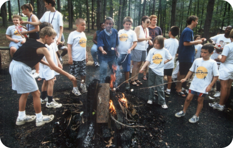 A colored photo, taken in the 1990s, showing a group of children at Camp Accomplish roasting marshmallows around a campfire.