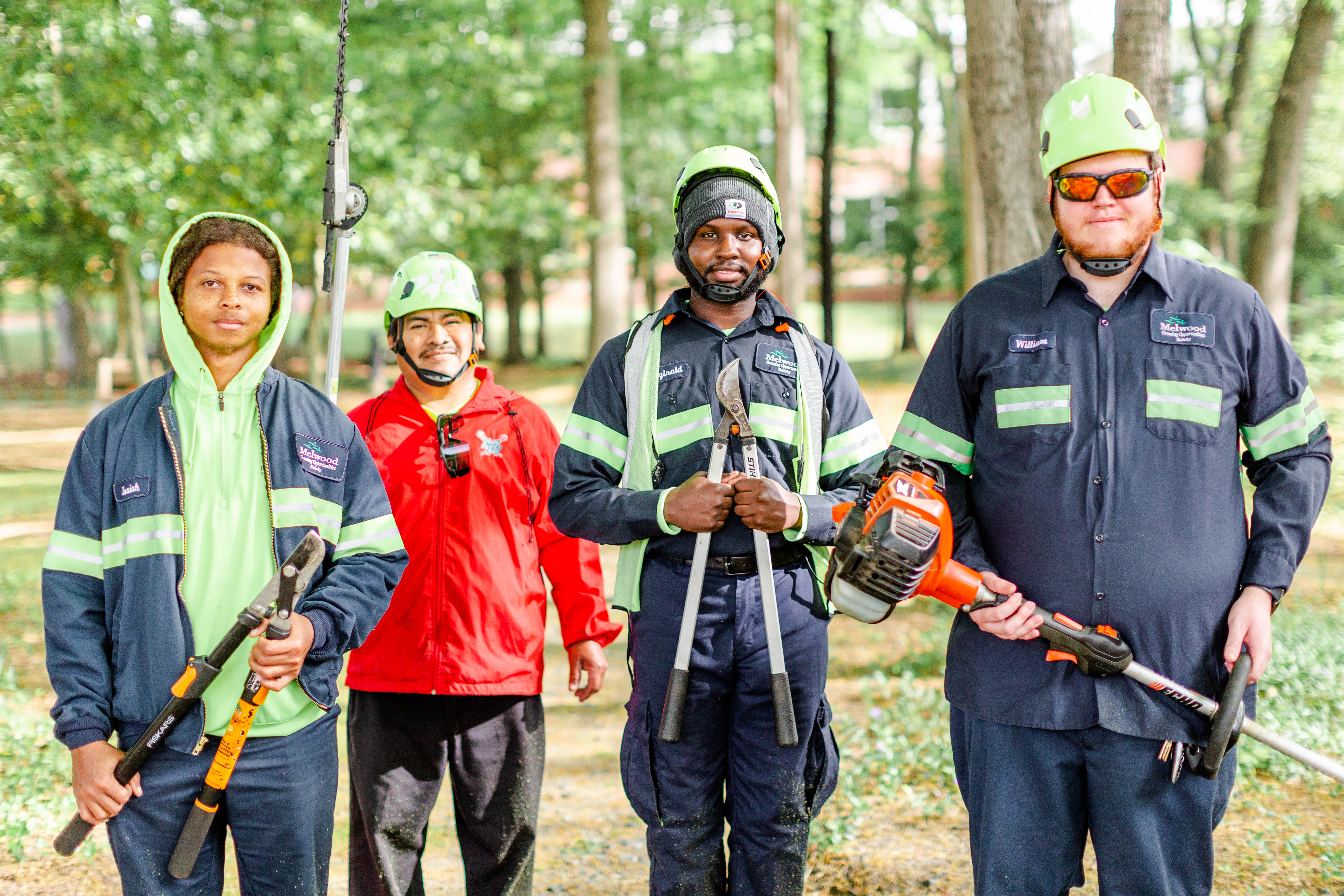 Group of four landscapers holding gear and wearing safety clothing, standing outdoors among trees