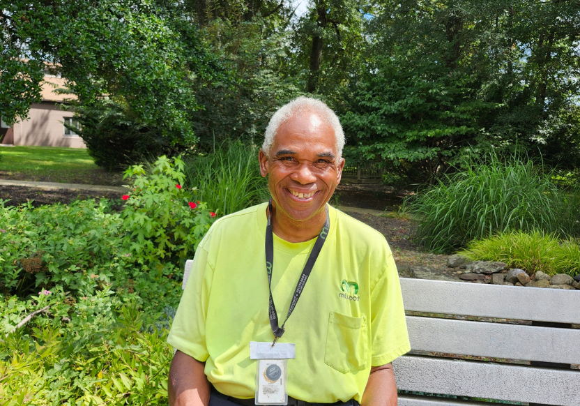 Lyndon Outlaw sits smiling on a bench at Melwood headquarters, wearing a yellow Melwood T-shirt and a badge.