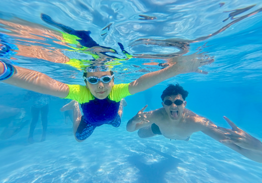 Camper and counselor underwater in a pool, both in goggles and with arms outstretched, at Camp Accomplish