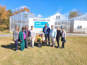 A group of people standing outdoors in front of greenhouses and a sign reading, "Welcome to Camp Accomplish"