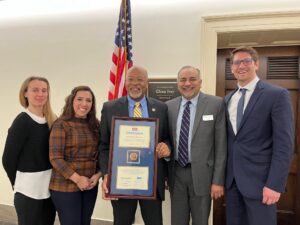 A group of people presenting Rep. Ivey with a plaque outside his office and in front of an American flag