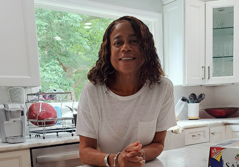 Delores Howard, smiling, in a white t-shirt in her kitchen, hands folded in front of her