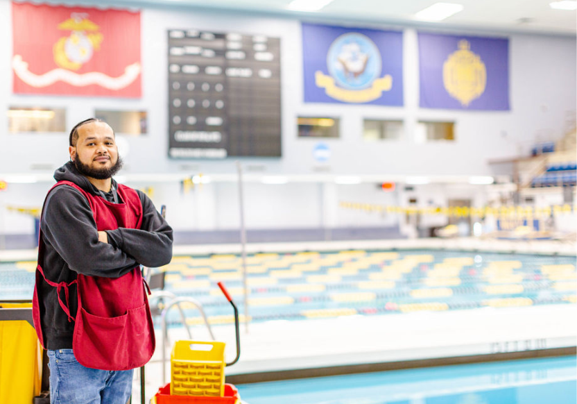 A man stands in front of an indoor pool. His arms are crossed and he's wearing a black hoodie with a red smock over it.