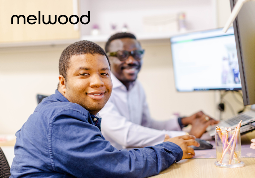 two people sitting at a computer typing, facing the camera and smiling; the word "Melwood" is emblazoned on the photo