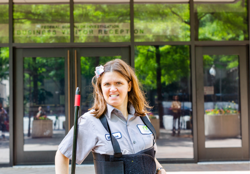 Woman in work gear, holding the handle of a broom or mop, stands smiling in front of glass doors