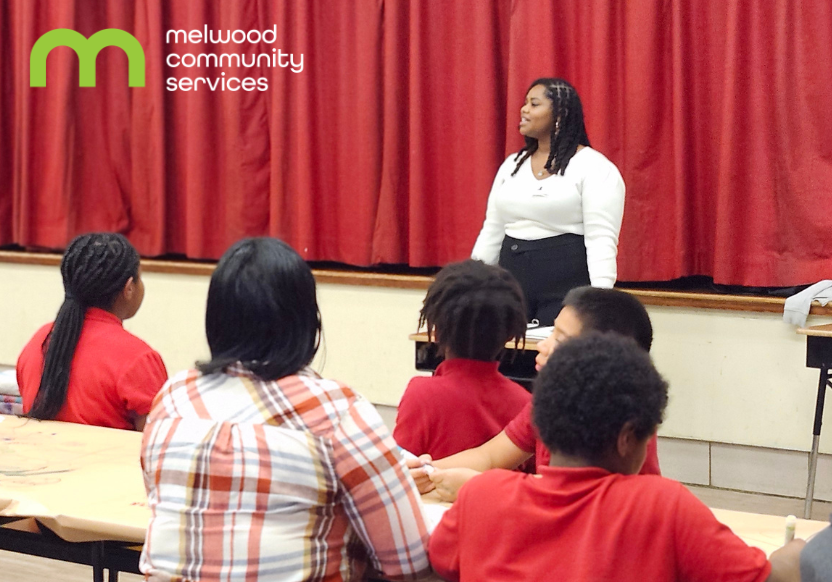 A woman in a white shirt stands before a red curtain on a stage, and in front of her are students at a table