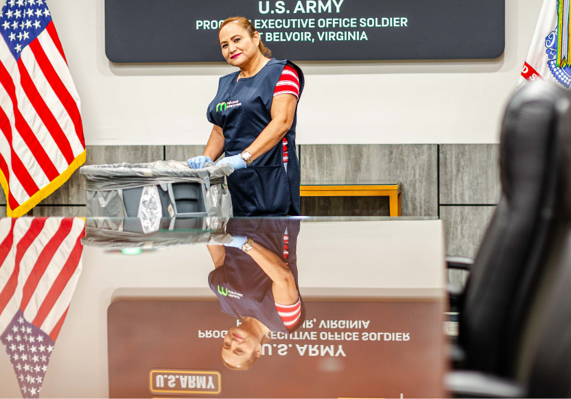 A woman in a Melwood Enterprises smock stands by an American flag and before a gleamingly clean tabletop. Behind her is a sign showing she is at a U.S. Army base at Fort Belvoir
