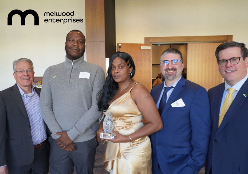 A woman in a gold dress holding an award flanked on either side by two men in business attire, with the words "Melwood Enterprises" emblazoned across the top left of the photo