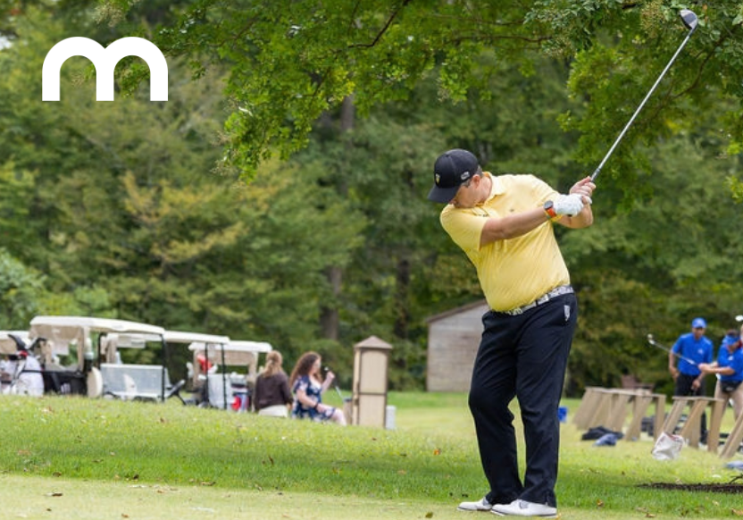 A man in a yellow shirt and black pants, golfing mid-swing, with golf carts and other golfers visible behind him
